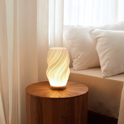 De Balaara Serene Wavecrest Lamp in een kalme slaapkamer, geplaatst op een houten tafel naast een bed met witte kussens, straalt zacht, warm licht uit dat een rustgevende sfeer creëert.